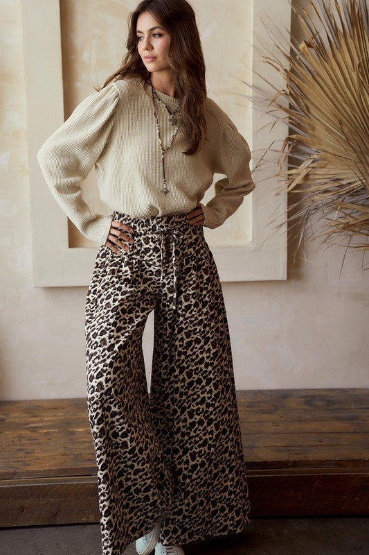 Front view — leopard print wide-leg pants with high rise and tie waist, flowy woven drape; styled with a beige knit sweater on model.