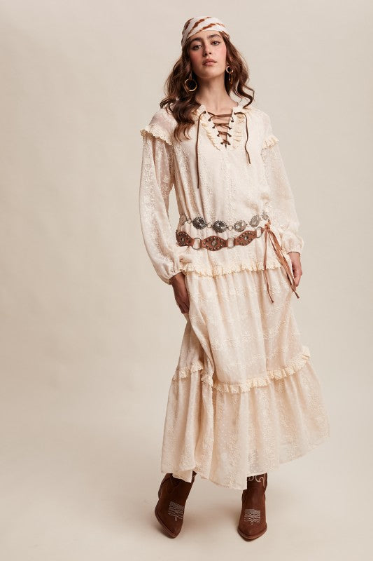 Model in ivory boho maxi dress with lace-up neckline, long puff sleeves, and tiered ruffle skirt, styled with concho belt, headscarf, and western boots.
