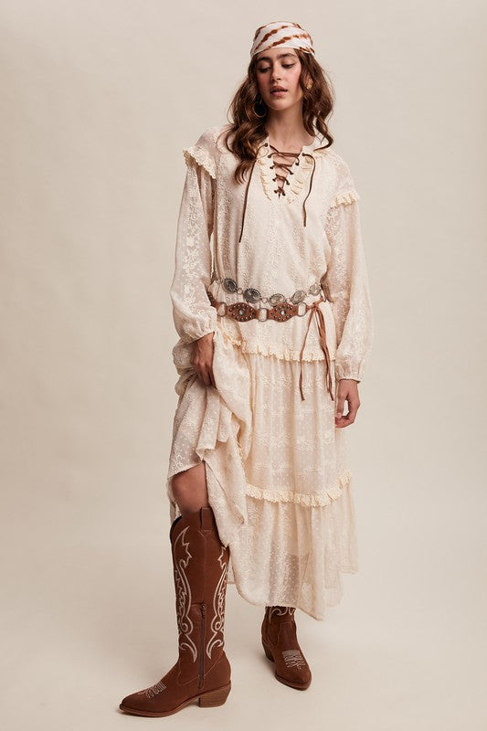 Model in ivory boho maxi dress with lace-up neckline, long puff sleeves, and tiered ruffle skirt, styled with concho belt, headscarf, and western boots.