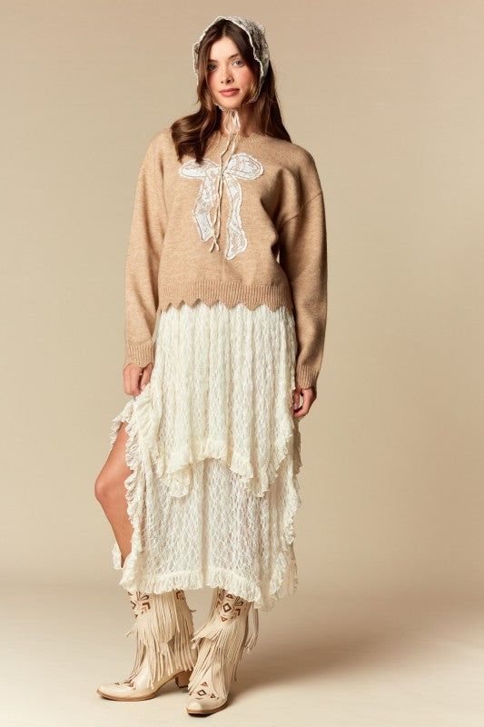 Camel knit sweater with white lace bow applique, relaxed fit, ribbed cuffs and scalloped hem, three-quarter front view on model.