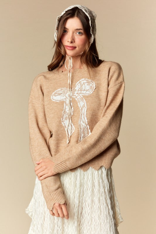 Camel knit sweater with white lace bow applique, relaxed fit, ribbed cuffs and scalloped hem, three-quarter front view on model.