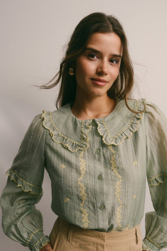 Model wearing sage green embroidered blouse with ruffled Peter Pan collar, button front, puff long sleeves and smocked cuffs, paired with tan trousers.