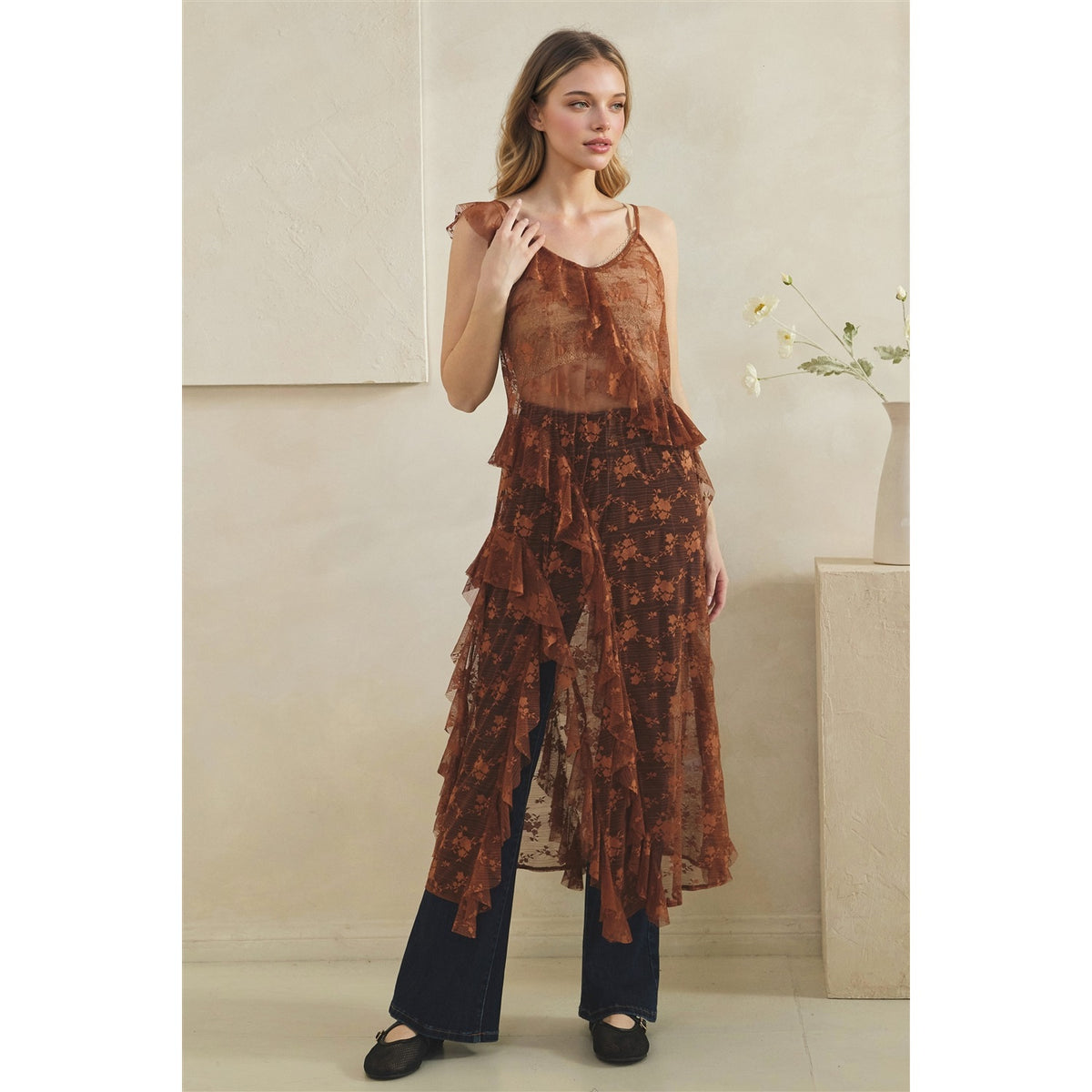 A woman models a copper sheer tulle top with a floral pattern and ruffled layers, paired with dark wide-leg jeans and black flats, standing in a minimalist beige room.