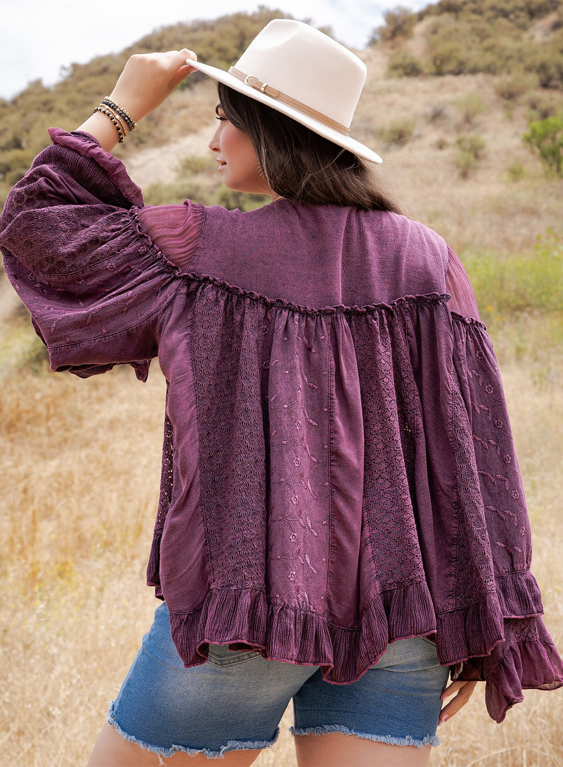 A woman wearing a purple textured bohemian-style blouse with ruffles, blue denim shorts, and a cream wide-brim hat, standing in a dry grassy outdoor field.