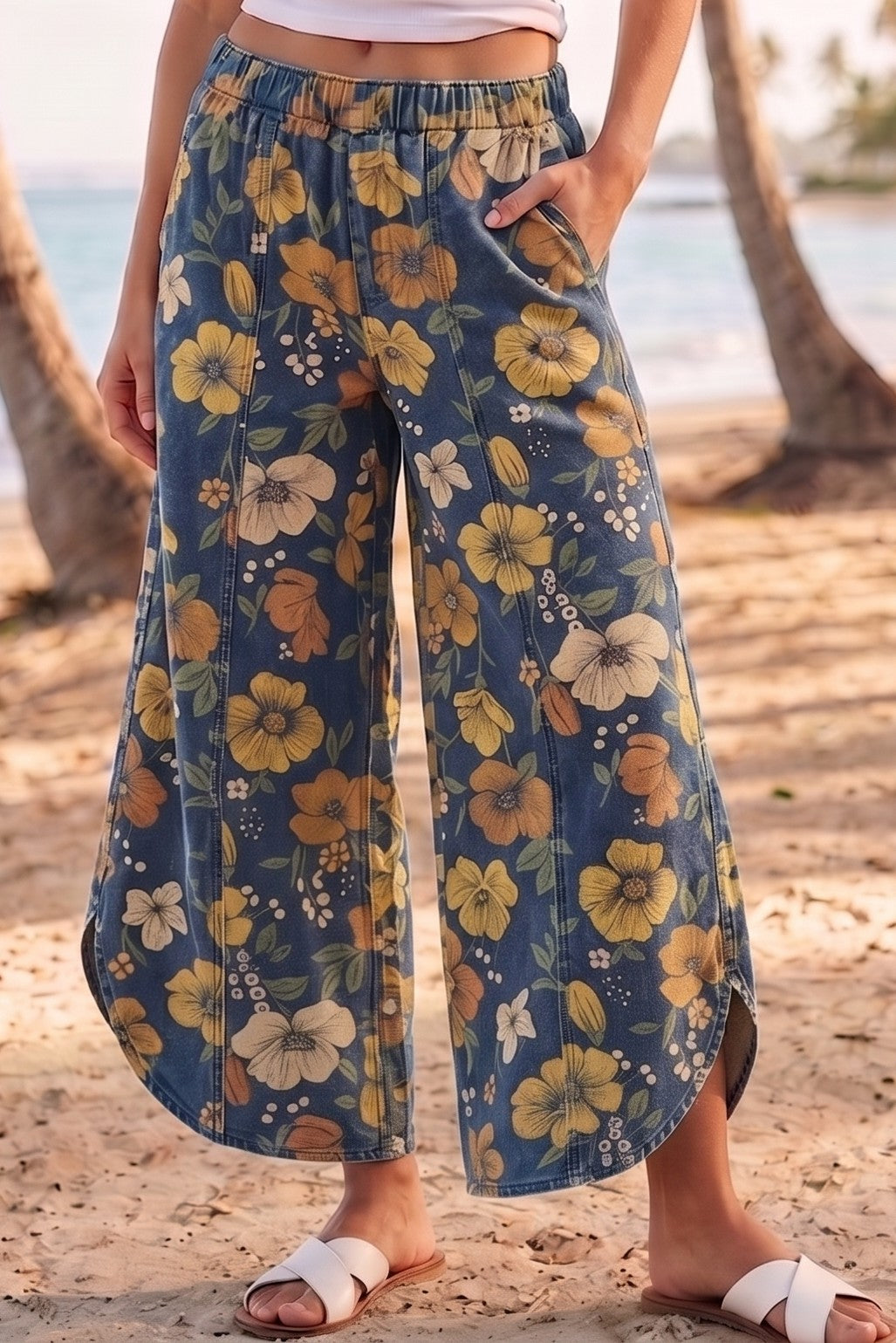 A person standing on a sandy beach wearing wide-leg cropped pants with a blue base and a yellow, orange, and white floral print. The person is also wearing white slide sandals.