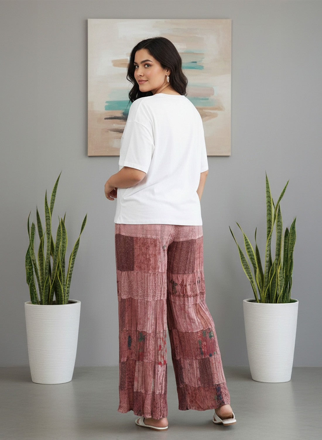 A woman stands with her back to the camera wearing a white t-shirt and dusty rose patchwork wide-leg trousers, posed between two potted snake plants against a gray wall with an abstract painting.