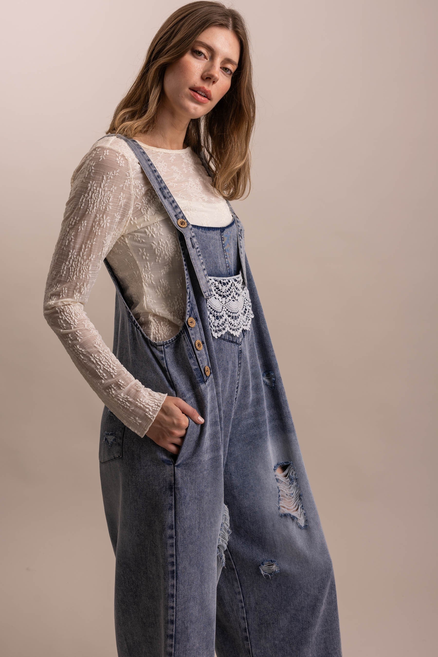Angled side view of distressed denim overall showing crochet lace bib pocket, button strap closures, and distressed leg detailing