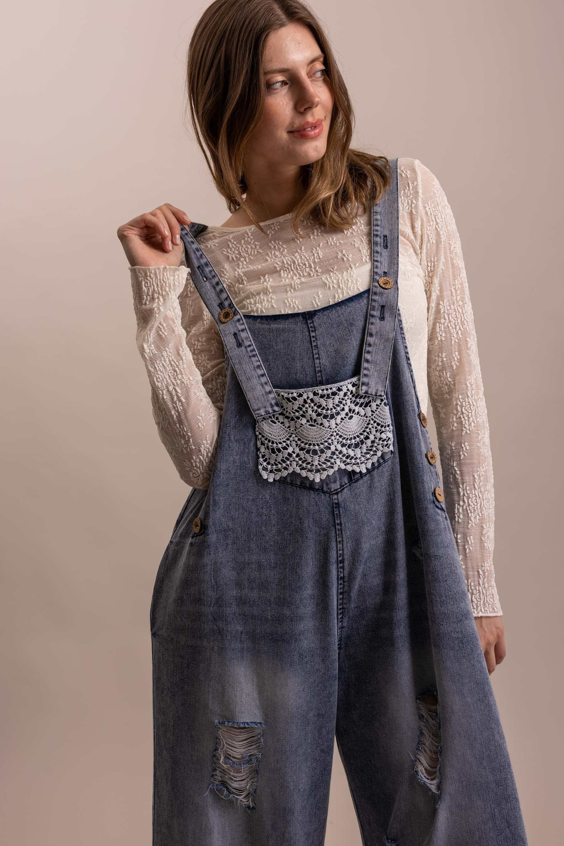 Model smiling in distressed wide leg denim overall with crochet lace bib pocket, tiered lace ruffle hem, and adjustable button straps