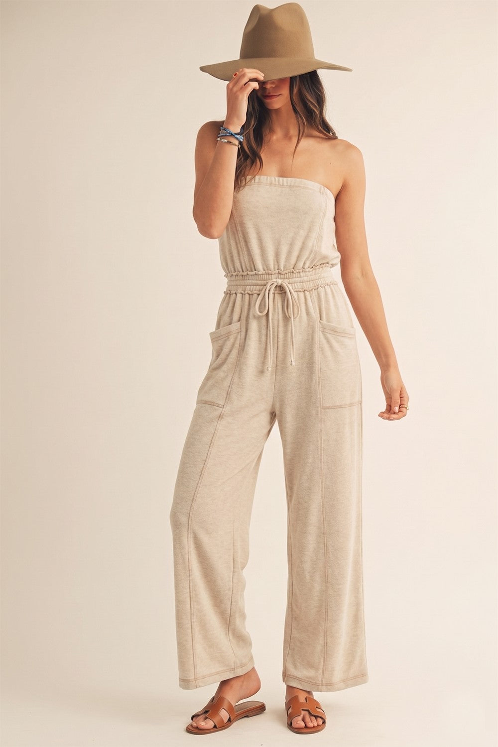 A woman modeling a beige strapless jumpsuit with a drawstring waist and wide legs, paired with a tan wide-brim hat and brown sandals, against a plain light beige studio background.