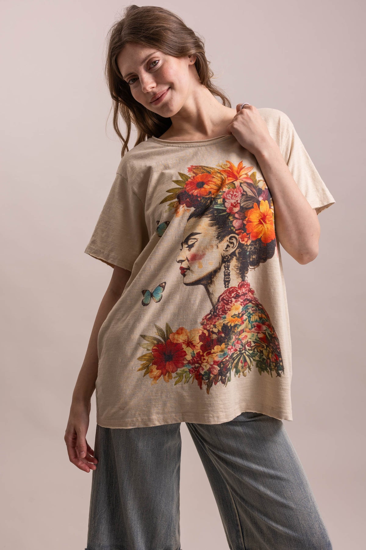 A woman wearing a beige graphic t-shirt with a colorful floral and portrait print, paired with wide-leg gray jeans, posing against a solid light beige background.