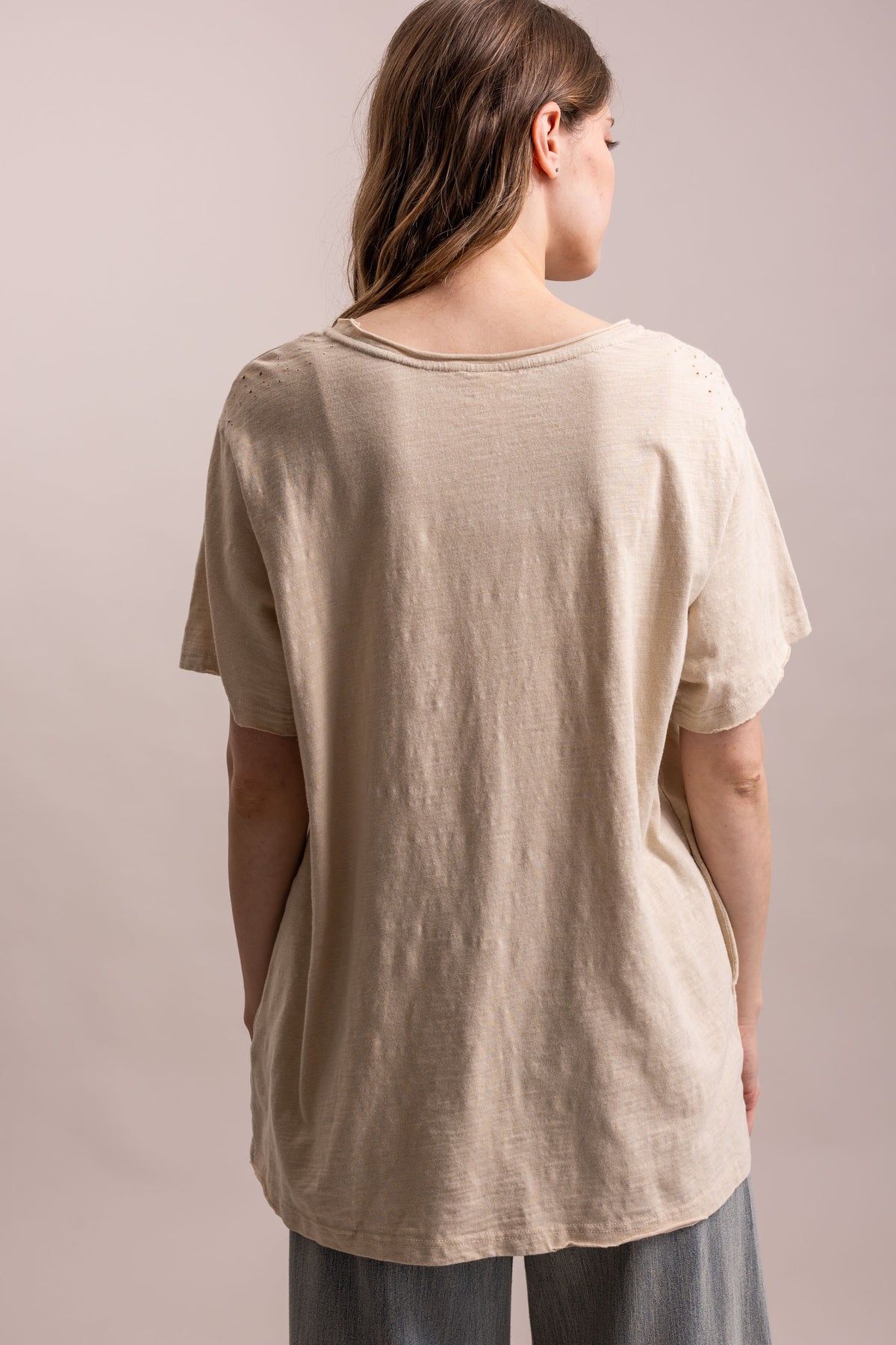 A woman viewed from behind wearing a loose, short-sleeve beige linen t-shirt and gray pants, set against a solid light beige studio background.