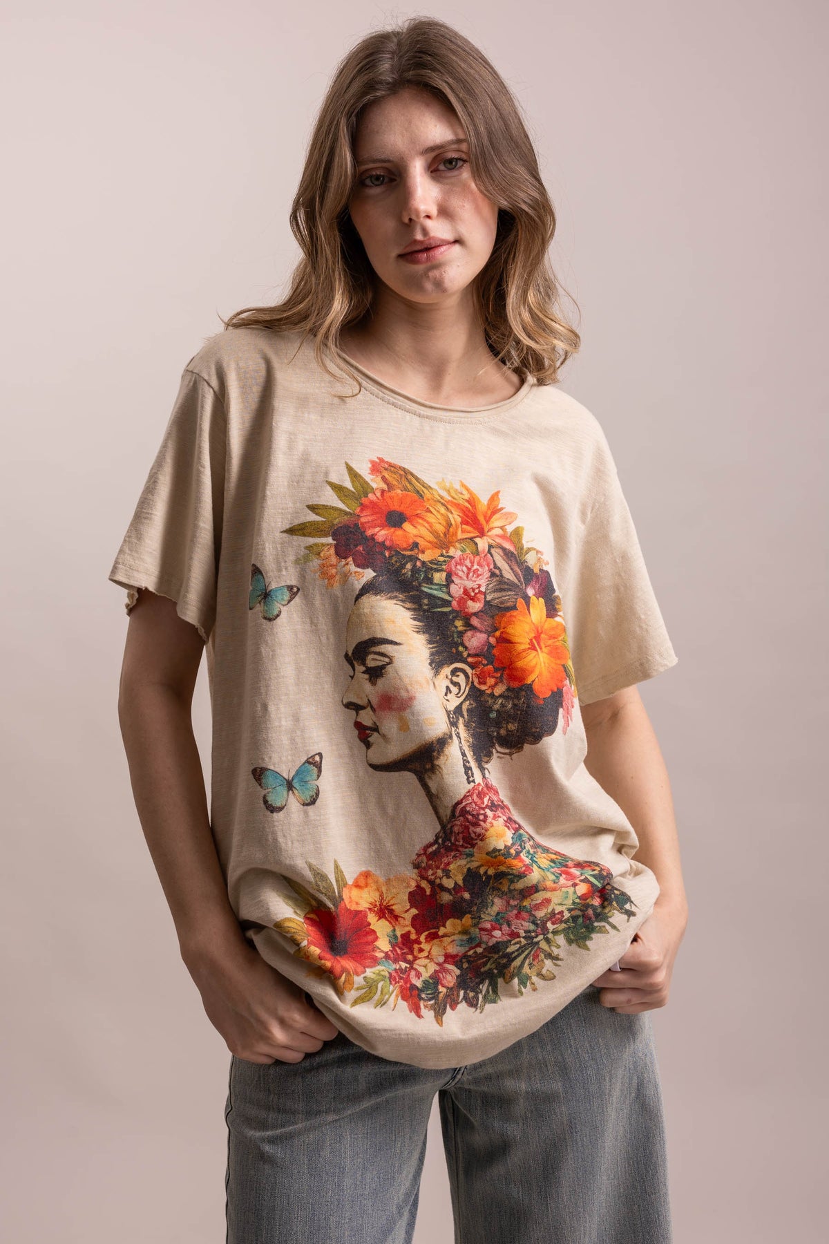 A woman modeling a beige short-sleeve t-shirt with a large graphic print of Frida Kahlo's profile adorned with vibrant orange and red flowers and blue butterflies, paired with light blue jeans against a solid light beige background.