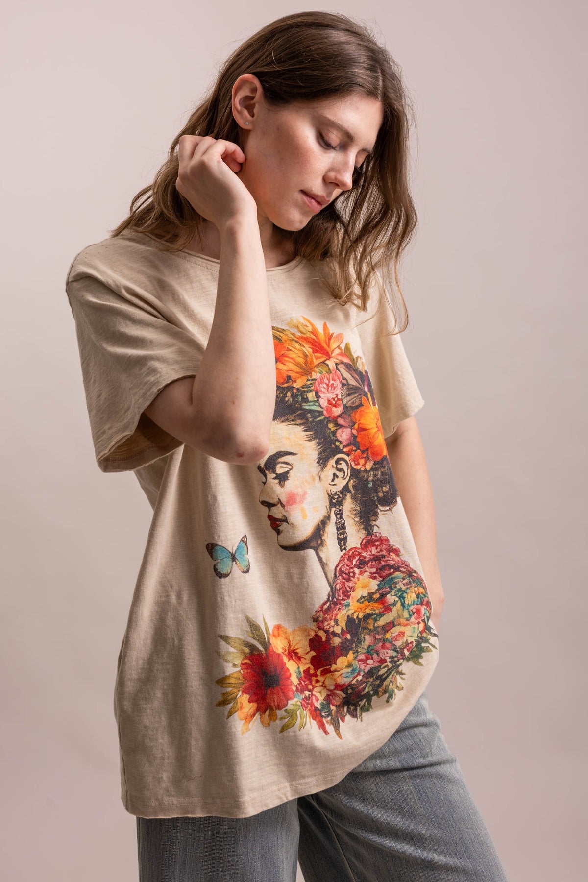 A woman wearing a beige oversized graphic t-shirt with a colorful Frida Kahlo-style floral portrait print, paired with gray jeans, against a solid light beige studio background.