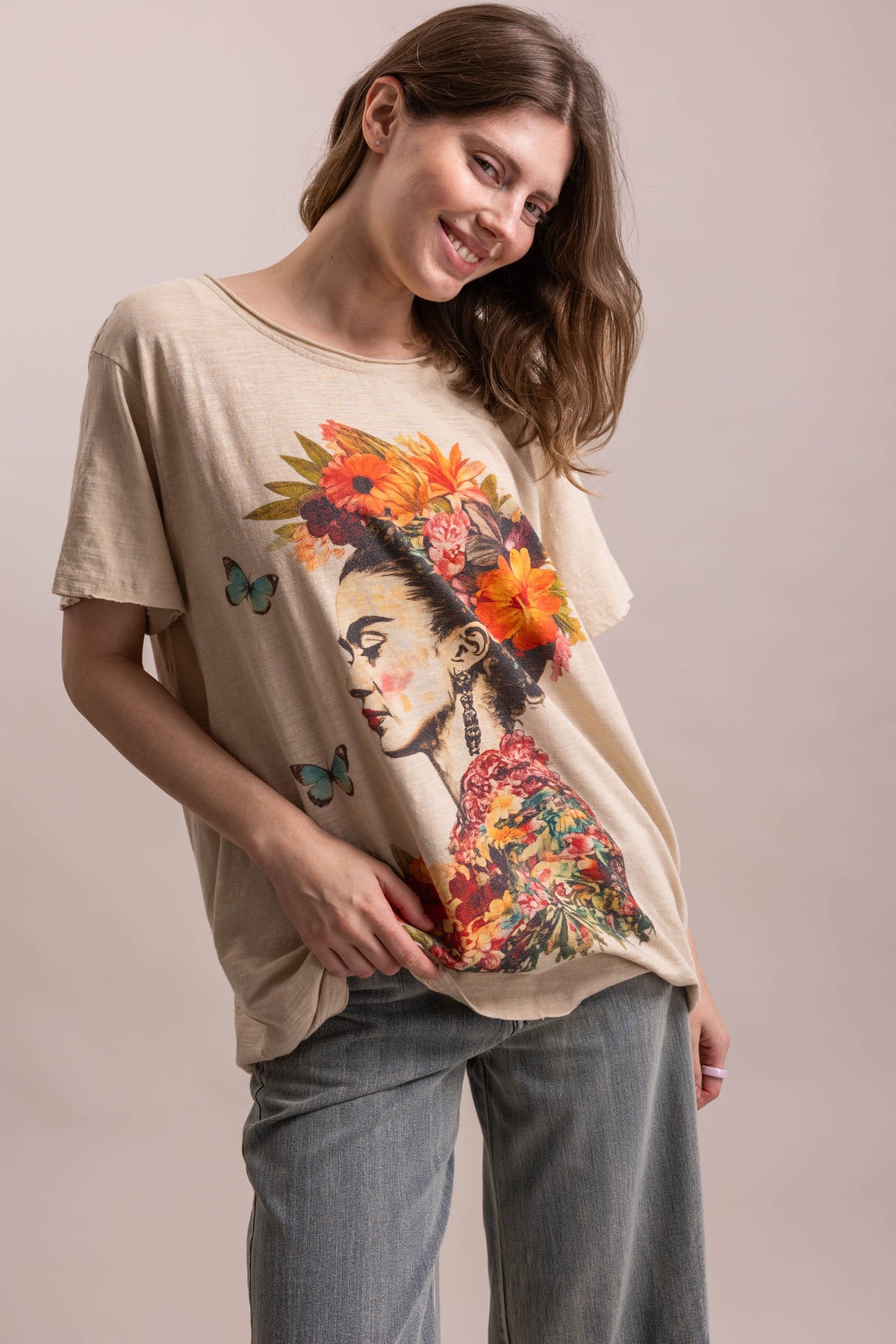 A woman wearing a beige oversized graphic t-shirt featuring Frida Kahlo's face surrounded by vibrant orange and yellow flowers and blue butterflies, paired with gray jeans, against a solid light beige background.