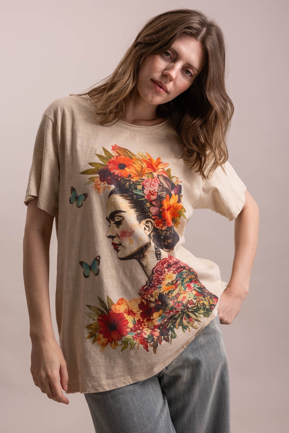 A woman wearing a beige oversized graphic t-shirt with a colorful Frida Kahlo-inspired floral portrait print, paired with light blue jeans, against a solid light beige background.