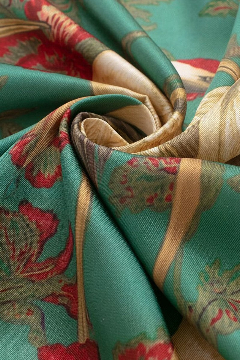 A close-up of a draped fabric with a teal green base, featuring a detailed floral and botanical pattern in red, beige, and olive green.