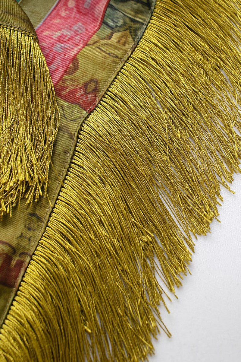 Close-up of a decorative fringe trim with dense metallic gold fringe over a patterned fabric backing in olive, pink, and gold tones, laid diagonally on a white background.
