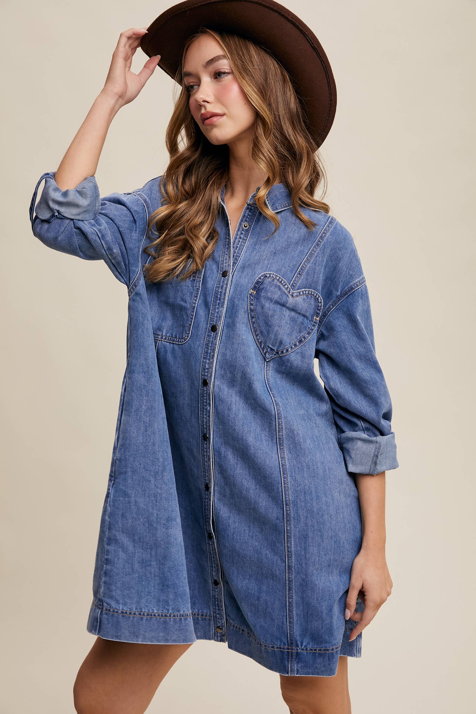 Medium-wash denim shirt dress with snap button front, heart-shaped chest pocket, rolled long sleeves, and a relaxed mini silhouette.