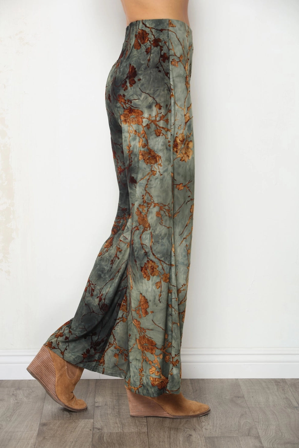 A person shown from the waist down wearing wide-leg pants with an abstract floral print in muted green, brown, and rust tones, paired with tan wedge sandals, standing on a wood floor against a white wall.