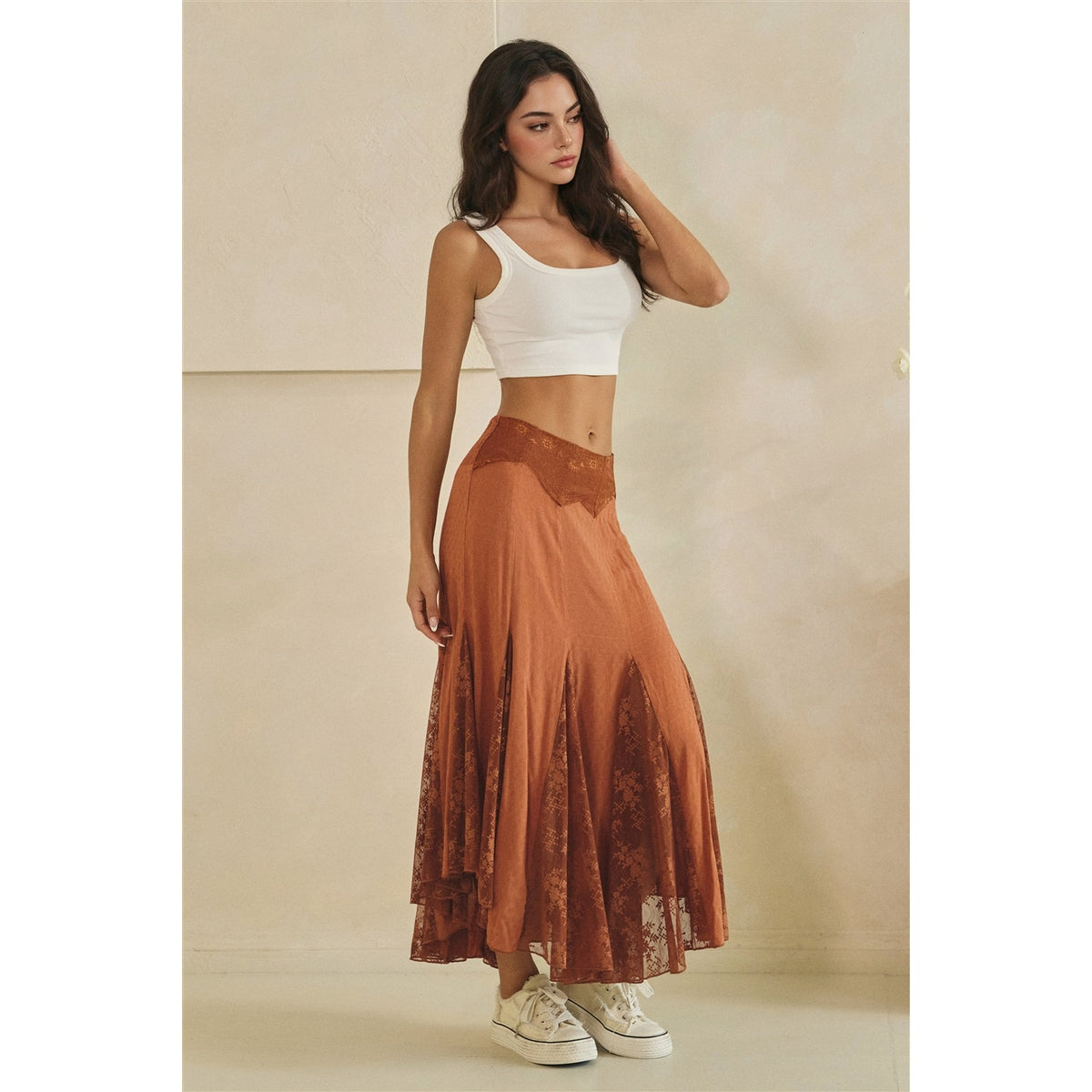 A woman modeling a rust-orange lace and mesh maxi skirt with a white crop top and white sneakers, standing against a neutral beige wall.