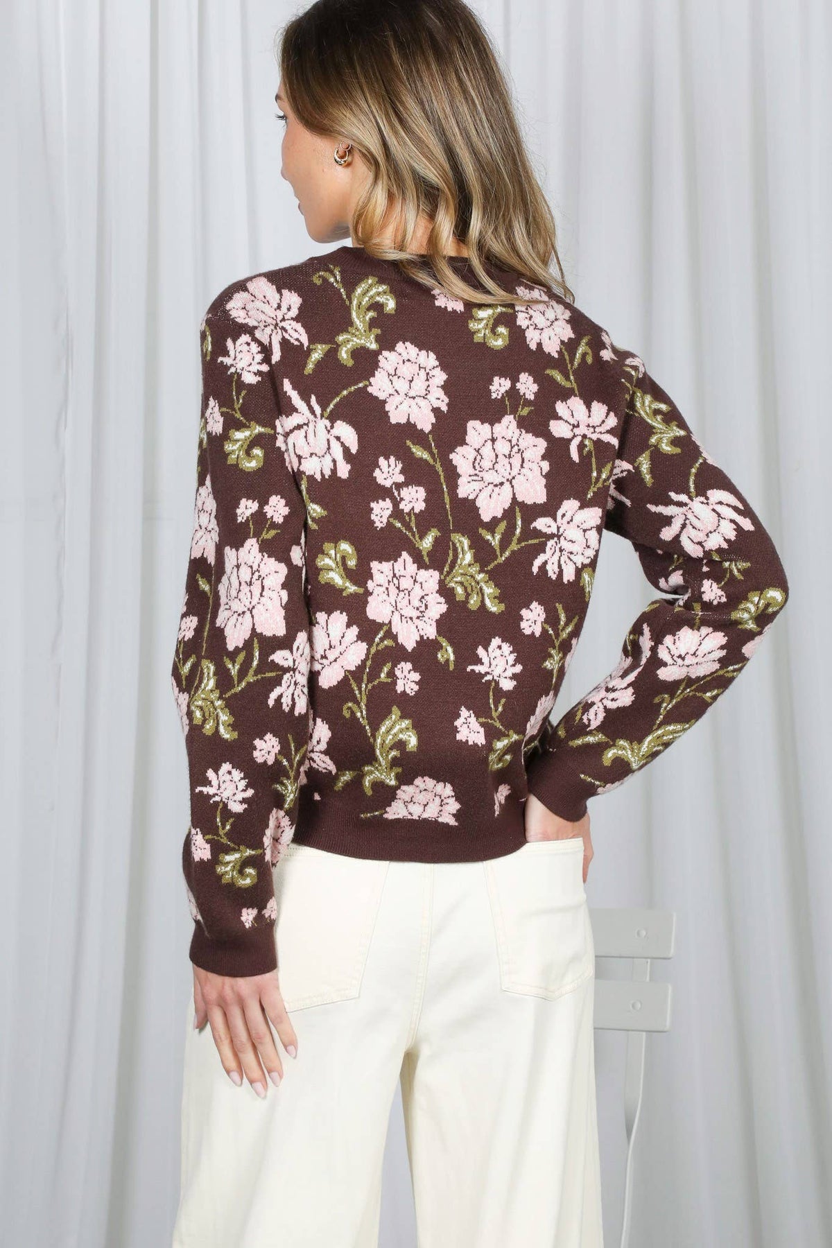 A woman shown from the back wearing a brown long-sleeve crewneck sweater with a pink and green floral pattern, paired with cream-colored pants, set against a white curtain background.
