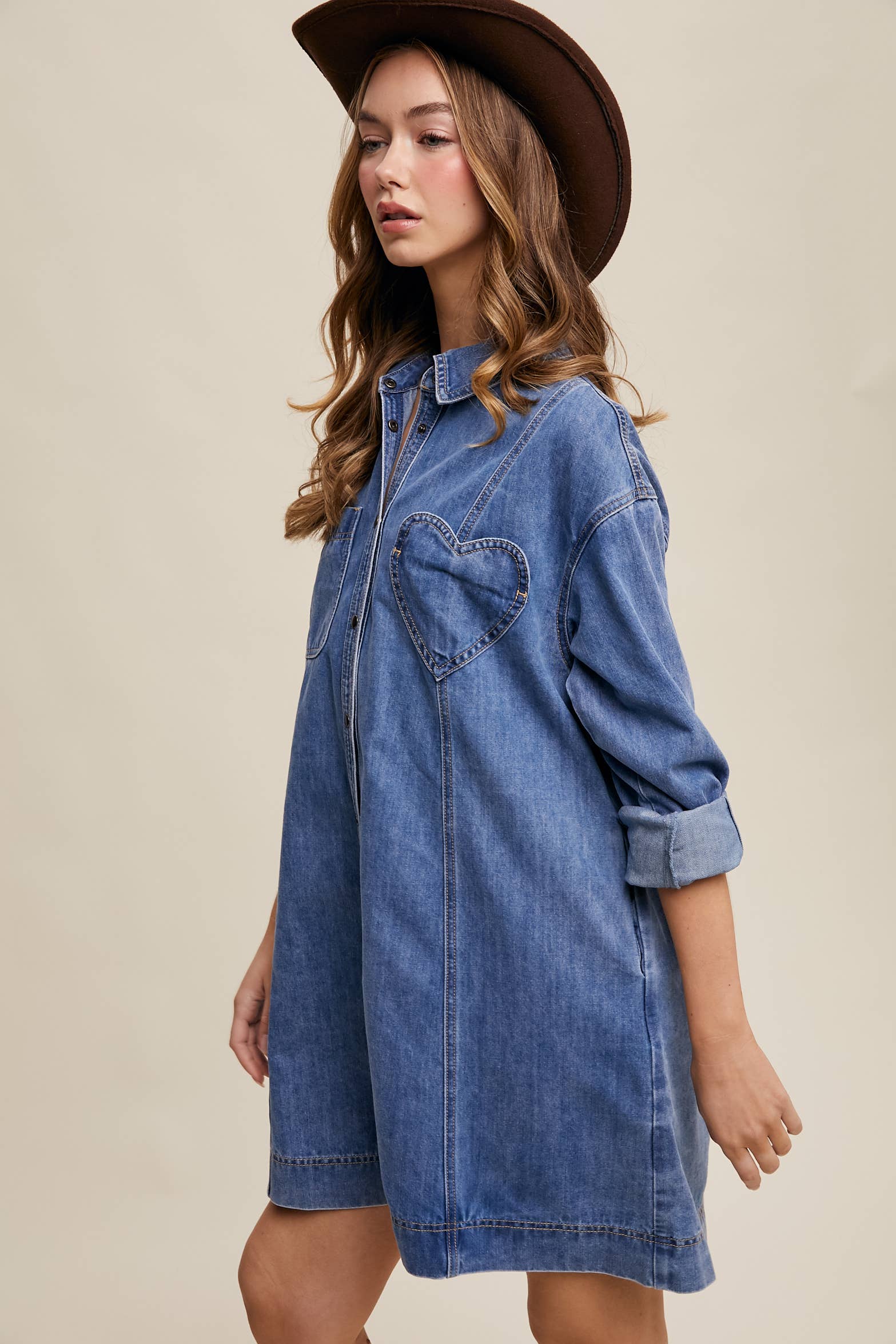 Side view- Medium-wash denim shirt dress with snap button front, heart-shaped chest pocket, rolled long sleeves, and a relaxed mini silhouette.