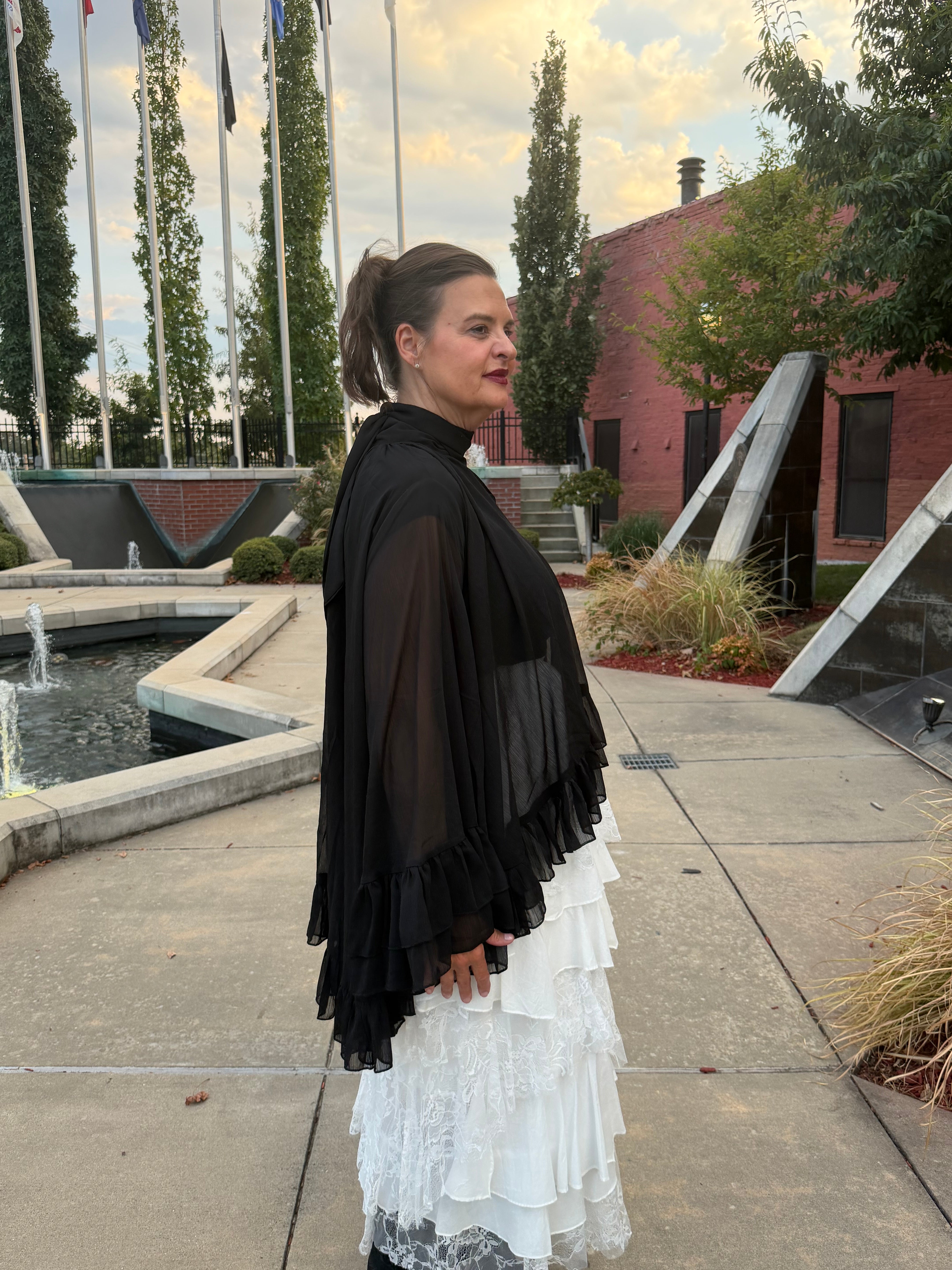 A woman standing outdoors in a courtyard, wearing a sheer black ruffled cape-style top over a white tiered skirt.