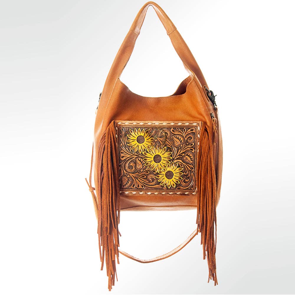 A tan leather hobo shoulder bag with long fringe, featuring a tooled floral panel on the front with three yellow sunflower designs, photographed against a light gray background.