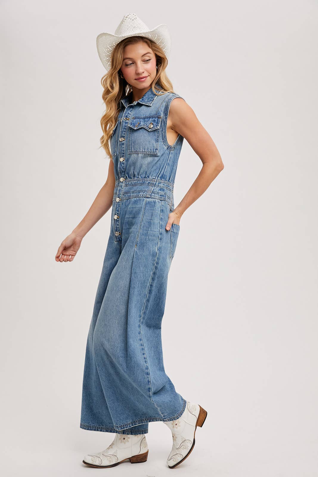 Side view of sleeveless denim jumpsuit with collared button-front and wide-leg legs, worn with brown slide sandals.