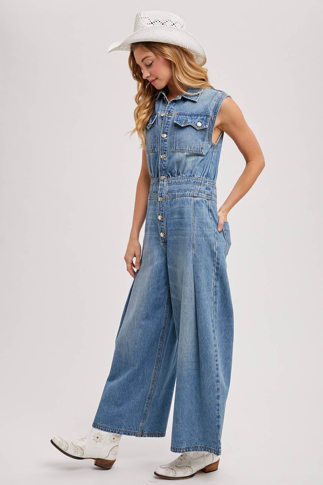 Side view of sleeveless denim jumpsuit with collared button-front and wide-leg legs, worn with brown slide sandals.