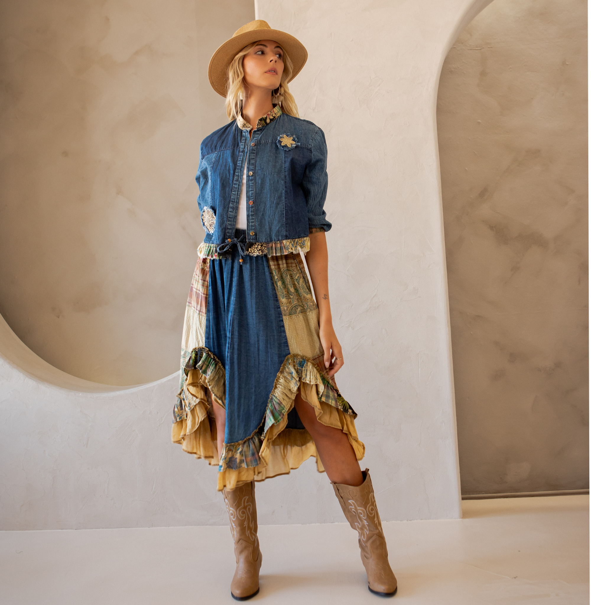 Model wearing denim patchwork jacket with frayed trim and floral appliqué, styled with a matching ruffled boho skirt and tan boots.