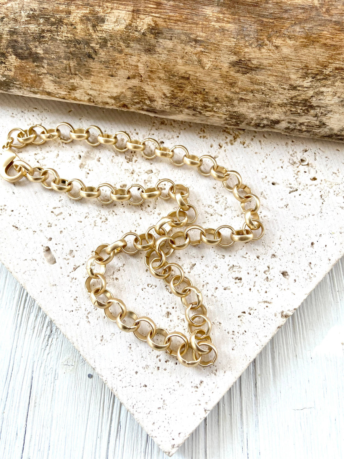 A gold chain necklace with interlocking circular links, draped across a textured white stone tile with a rustic wooden log in the background.
