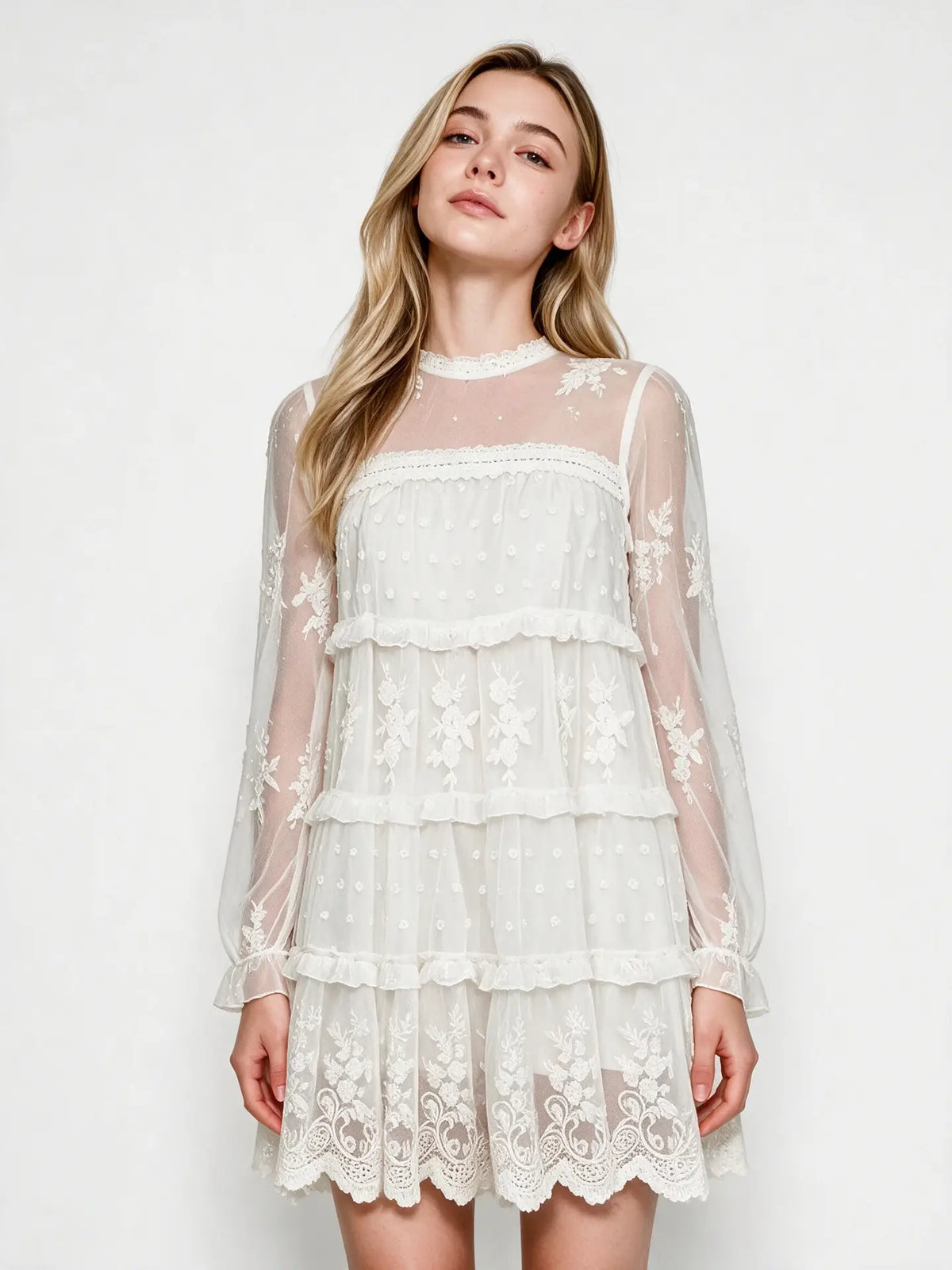 Close front view of the white lace dress showing floral embroidery, a sheer mesh yoke, crochet neckline trim, and ruffled tiers.
