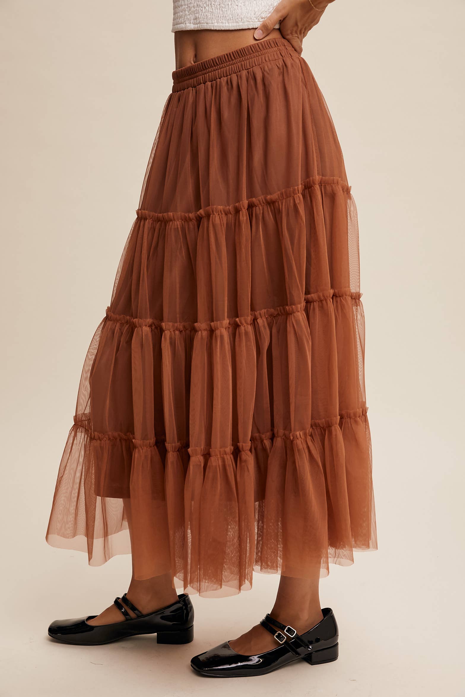 Side view — rust tulle tiered midi skirt with elastic waist and ruffle tiers, sheer overlay; model in black Mary Jane flats.