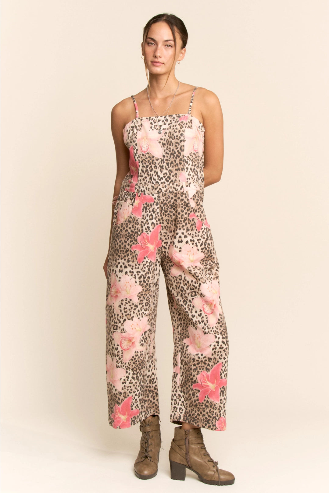 A woman modeling a sleeveless wide-leg jumpsuit with a leopard print base and pink floral pattern, paired with brown ankle boots, against a solid beige studio background.