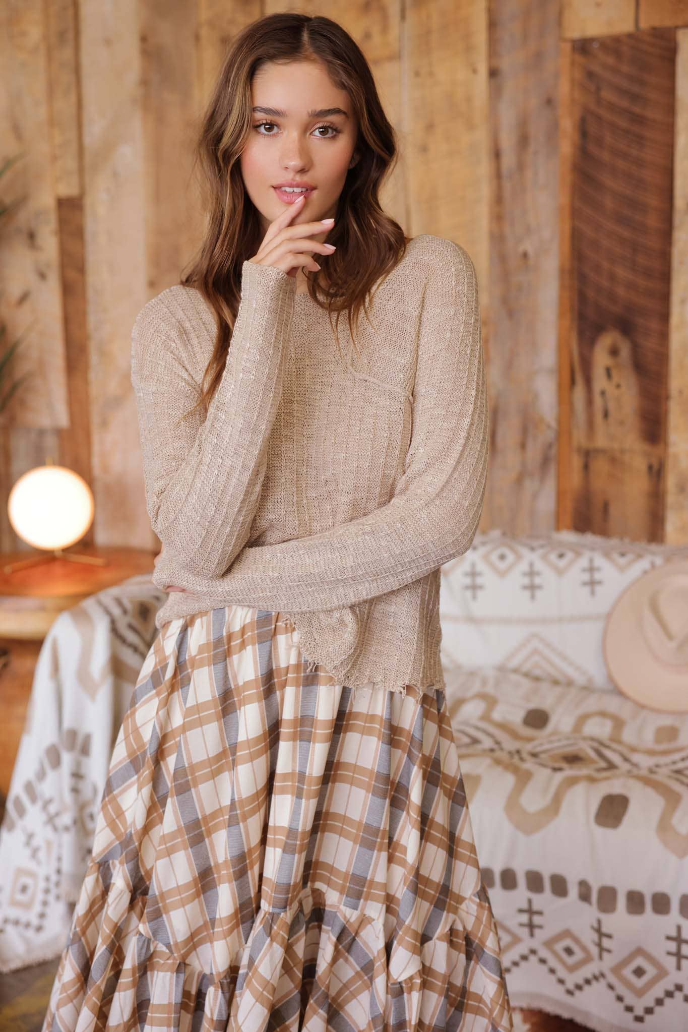 Model wearing taupe distressed lightweight distressed sweater with ribbed knit texture and front pocket, paired with a tan plaid ruffle skirt and sandals