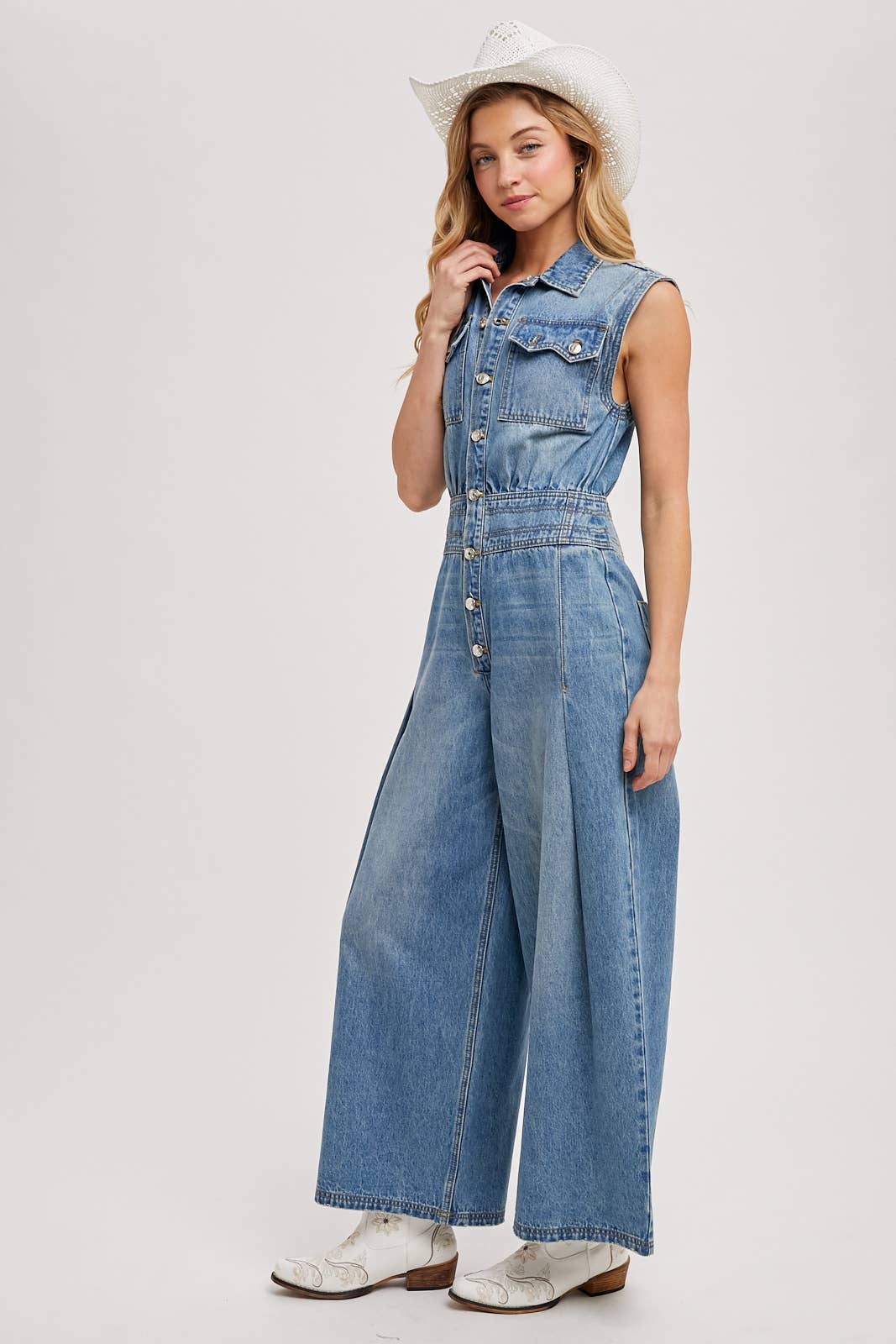 Side view of sleeveless denim jumpsuit with collared button-front and wide-leg legs, worn with brown slide sandals.