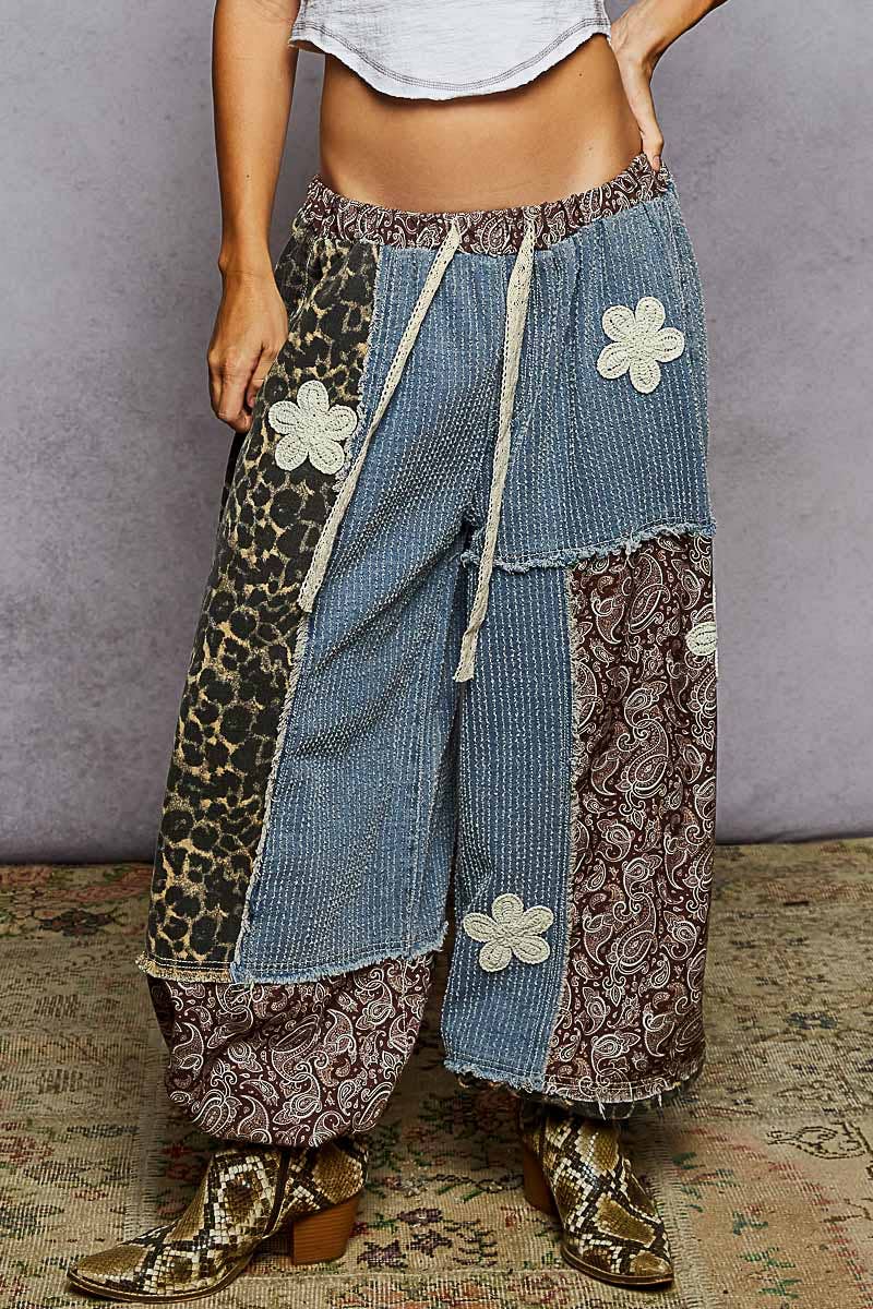 A person wearing loose patchwork harem-style pants with blue denim, leopard print, paisley, and floral applique, paired with snakeskin boots against a gray wall and patterned rug.