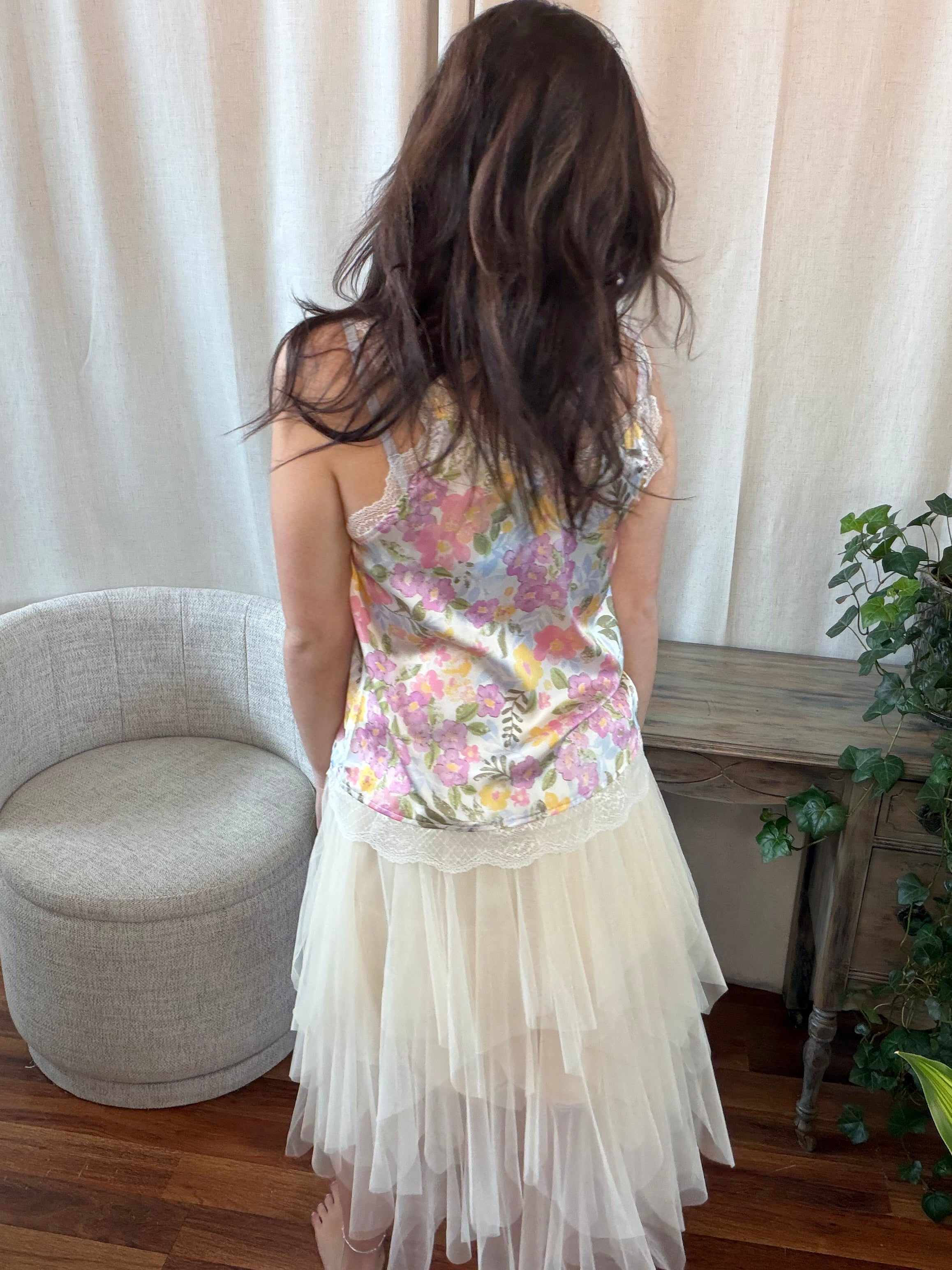 Woman wearing a floral top and white skirt in a room with curtains and a chair.