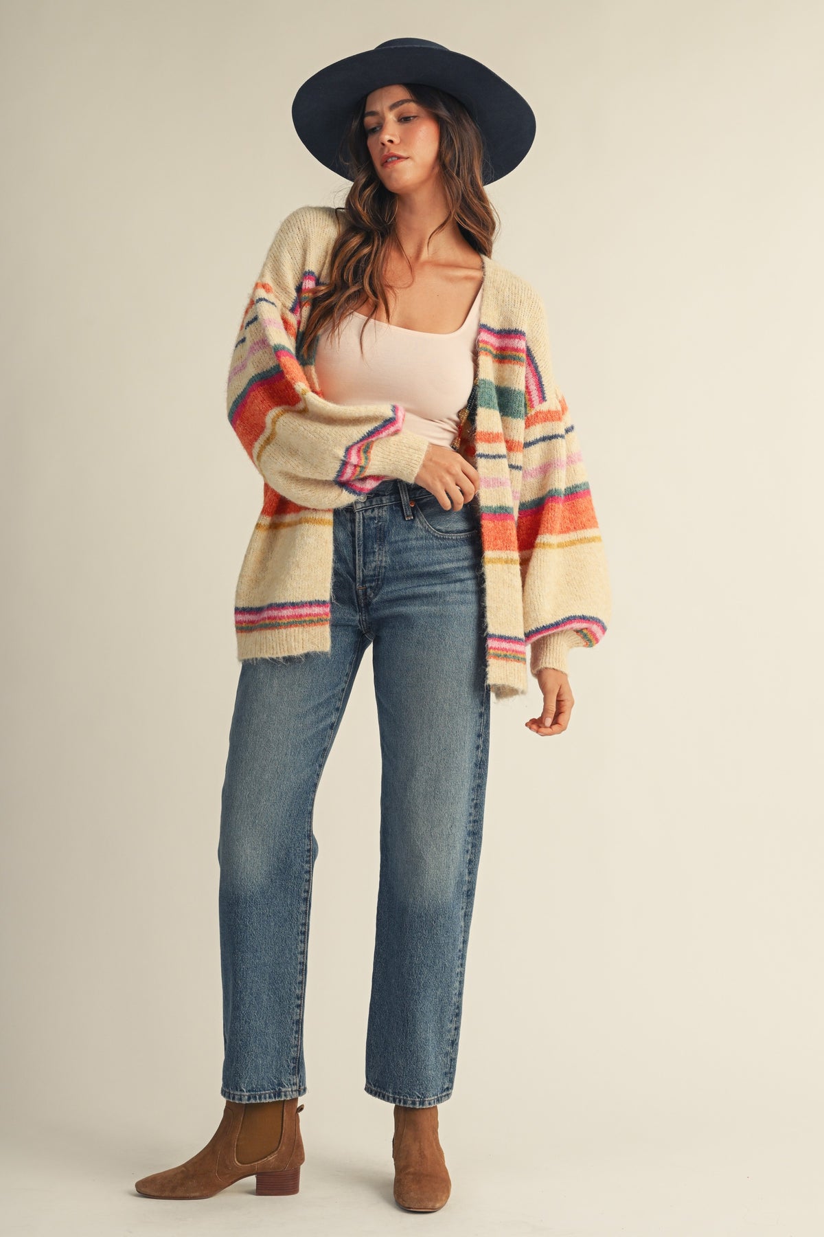 A woman modeling a cream knit cardigan with colorful horizontal stripes in red, orange, green, and blue, styled with a black wide-brim hat, a light pink tank top, blue jeans, and brown ankle boots against a solid beige studio background.