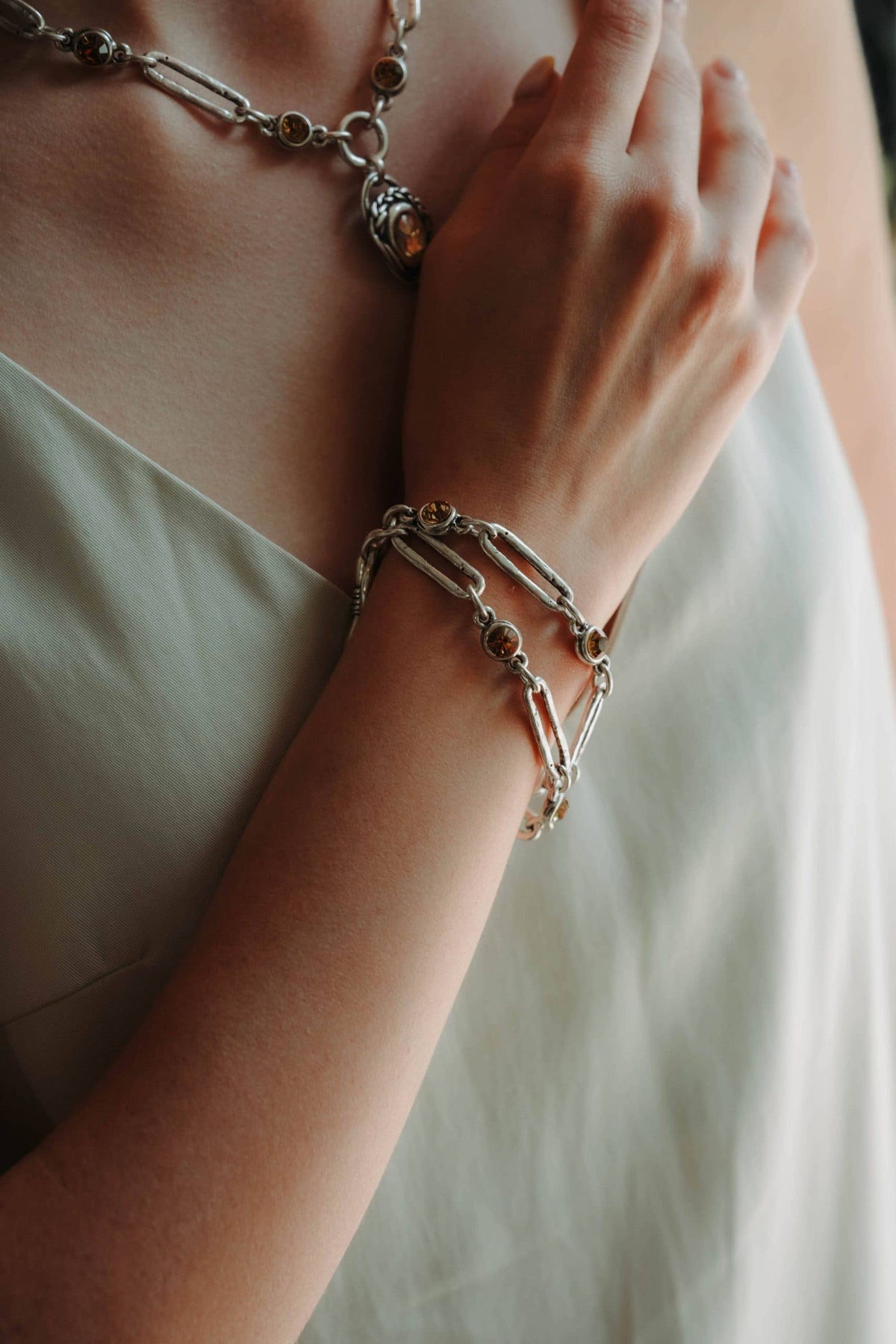 A close-up lifestyle shot of a person wearing a layered stack of silver chain bracelets on one wrist, along with a matching silver chain necklace. The person is wearing a light-colored, off-white satin dress.