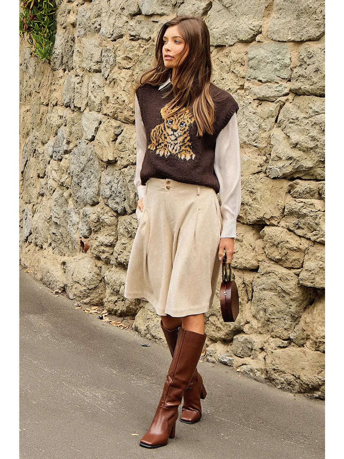Full-length view of a brown leopard cub sweater vest styled with a cream blouse, beige shorts, tall brown boots, and a handbag.
