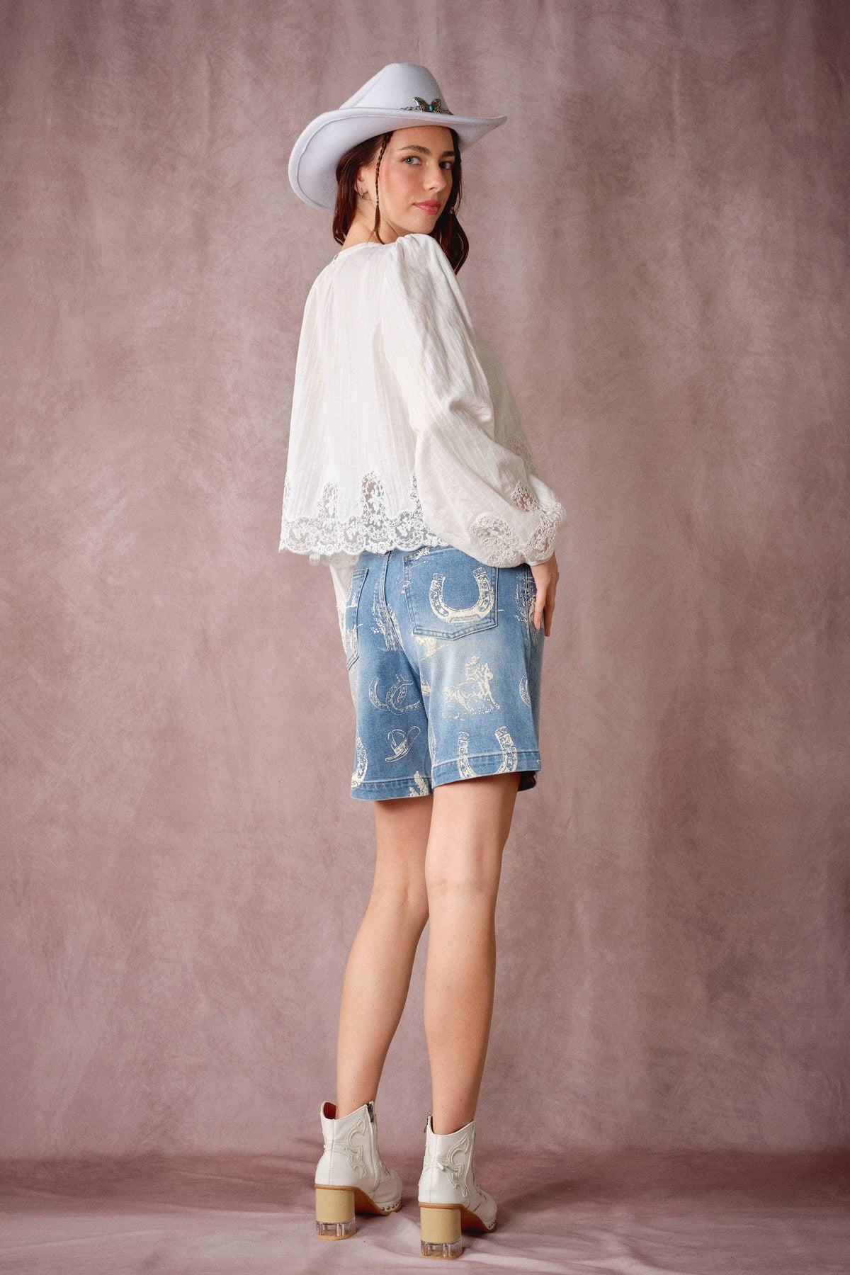 A woman modeling a white long-sleeve blouse with lace trim, light-wash denim shorts with western embroidery, and white cowboy boots, posed against a textured mauve studio backdrop.