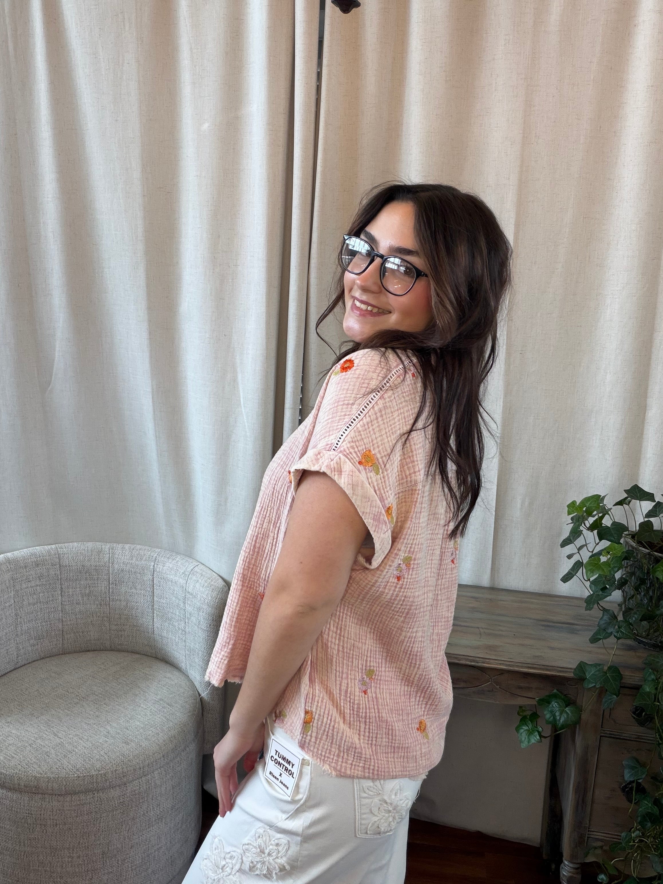 Woman wearing a pink blouse with floral patterns in a room with curtains and a chair.