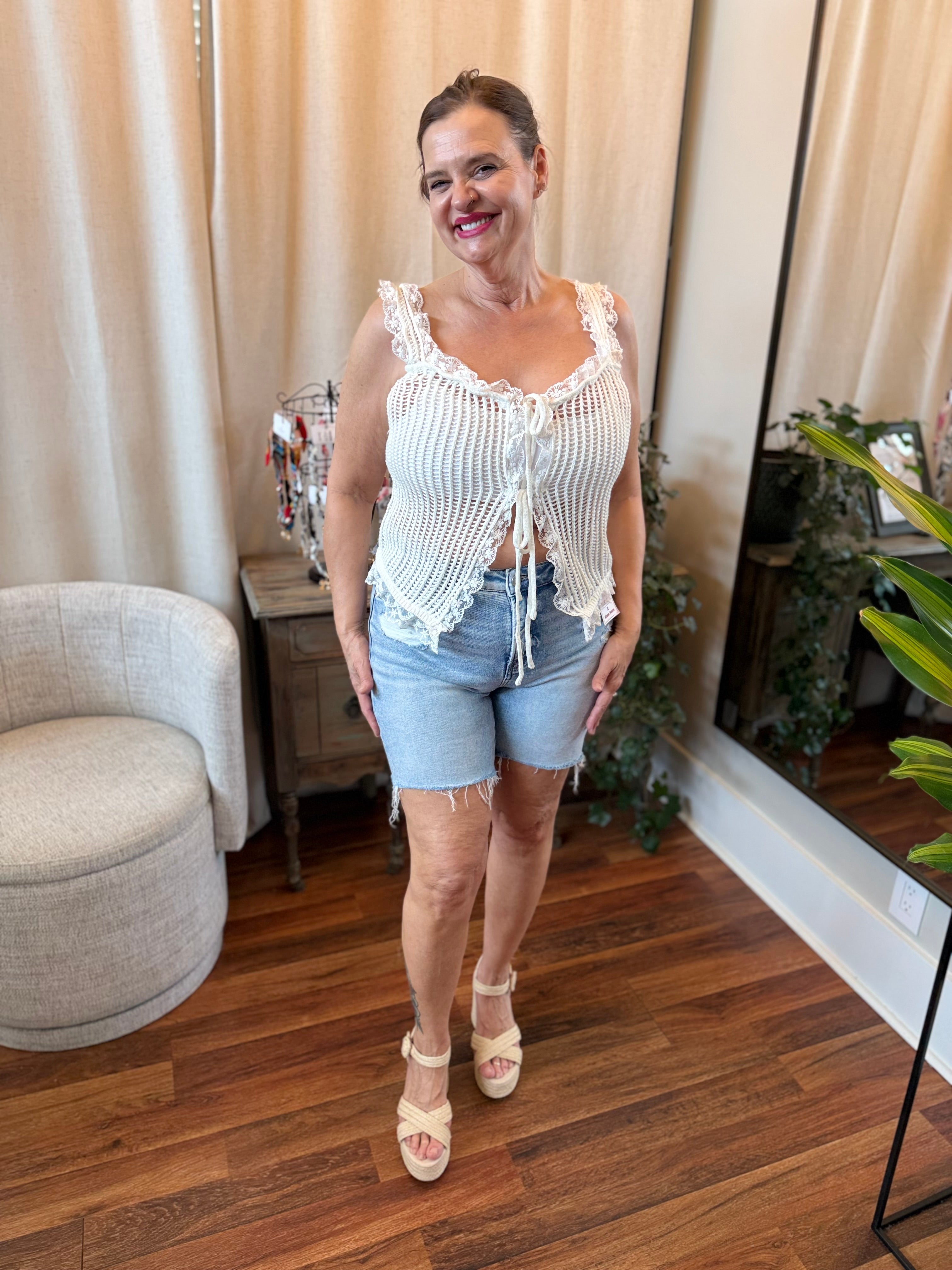 Woman in a white lace top and denim shorts standing in a room with curtains and plants.