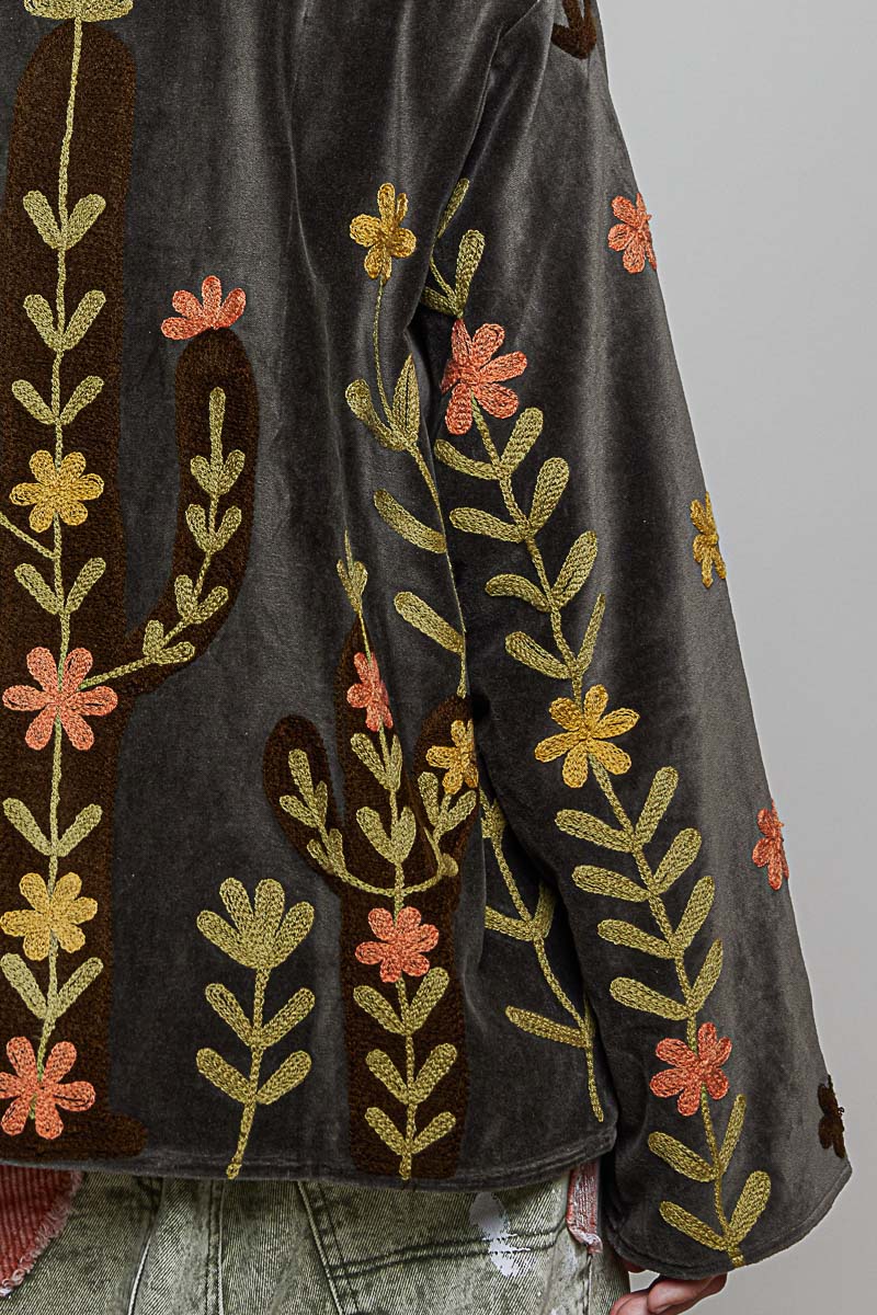 Close-up of a dark gray velvet garment with embroidered cactus and floral motifs in olive green, mustard yellow, and coral pink.