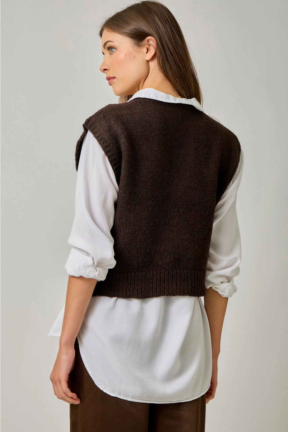 Back view of the brown sweater vest, showing its plain knit back and ribbed hem layered over a white collared shirt.

