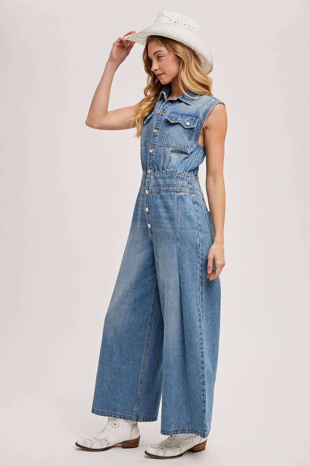 Side view of sleeveless denim jumpsuit with collared button-front and wide-leg legs, worn with brown slide sandals.