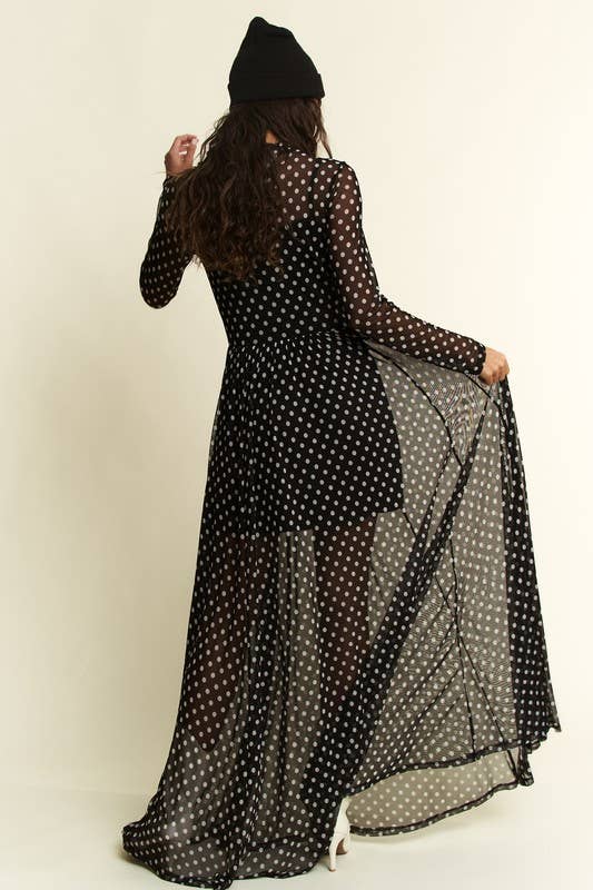Back view of a black polka-dot mesh long-sleeve maxi dress with a sheer overlay and short slip; model in a black beanie lifts the flowy skirt.