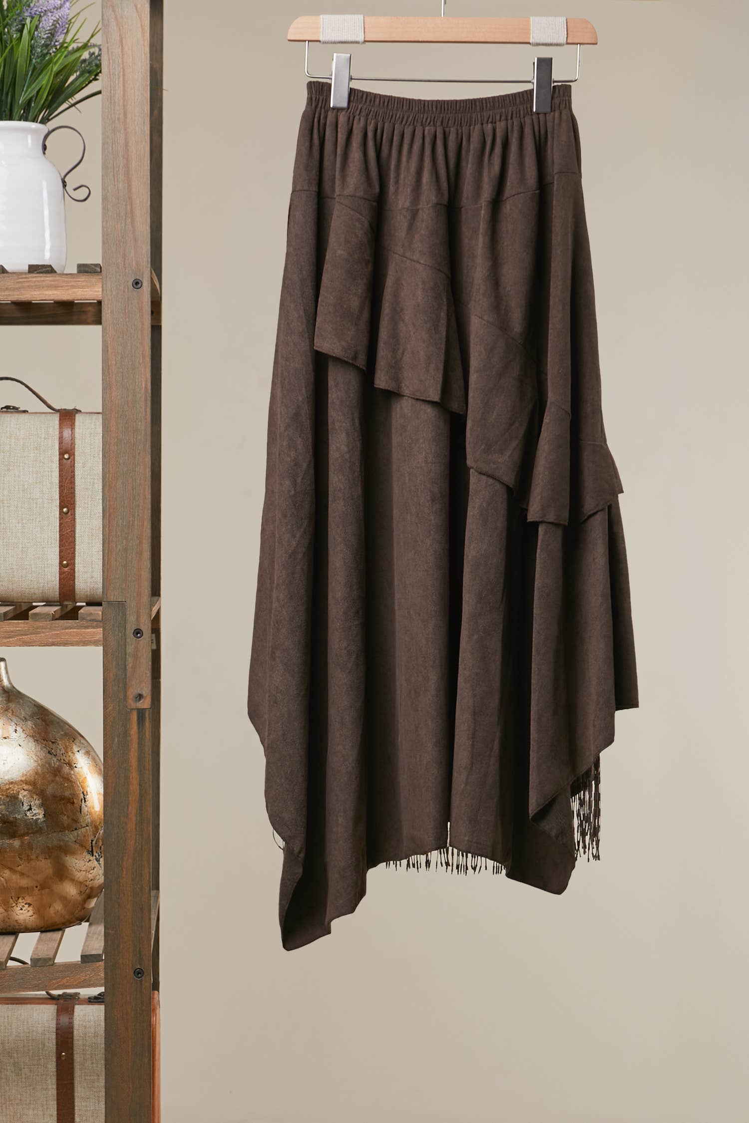 A dark brown tiered midi skirt with a textured, suede-like finish, hanging on a wooden hanger against a beige wall next to a rustic wooden shelf.